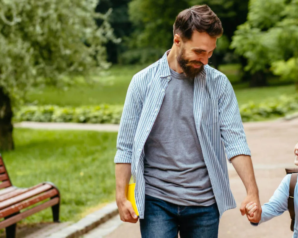 A smiling man and child hold hands, walking on a park path. The man carries a yellow folder. Lush greenery and a wooden bench frame the cheerful scene.