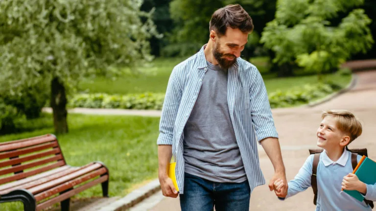 A smiling man and child hold hands, walking on a park path. The man carries a yellow folder. Lush greenery and a wooden bench frame the cheerful scene.