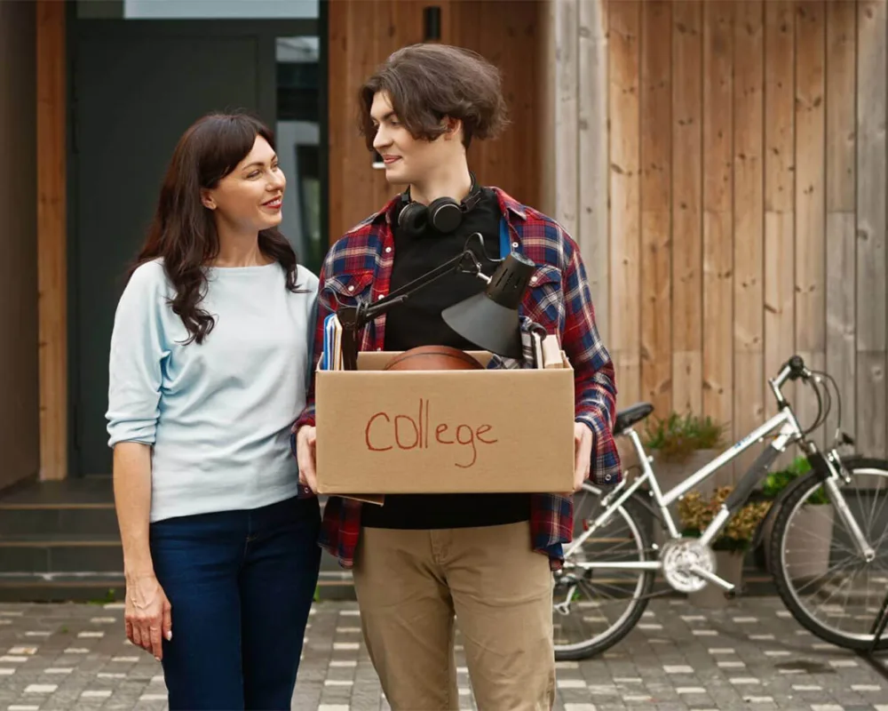 Portrait of adult pretty woman and young boy holding box labeled "College" standing outside and looking at eachother with smile.