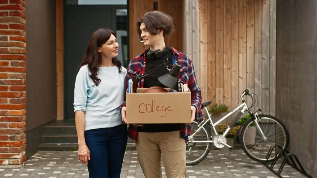 Portrait of adult pretty woman and young boy holding box labeled "College" standing outside and looking at eachother with smile.