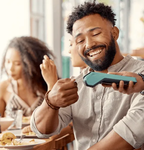A man smiles warmly while paying with a card reader in a lively cafe. Two women are chatting in the background, creating a joyful, relaxed atmosphere.