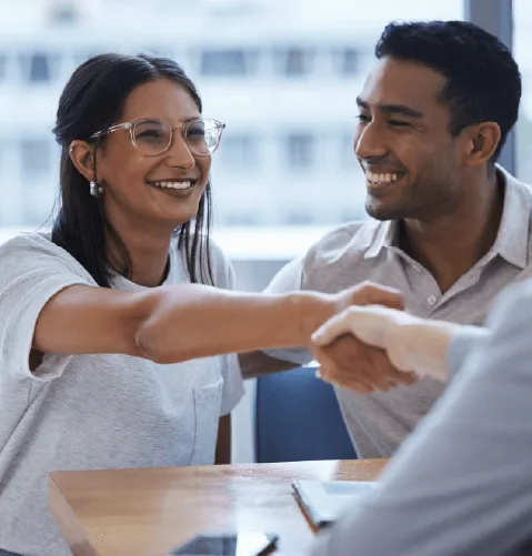 A smiling woman in glasses confidently shakes hands with someone off-frame, sitting beside a smiling man. They appear relaxed and positive in a light-filled office.