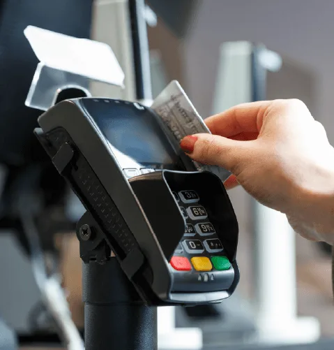 A hand with red nail polish swipes a credit card through a card reader at a checkout counter. Background is blurred, suggesting a retail setting.