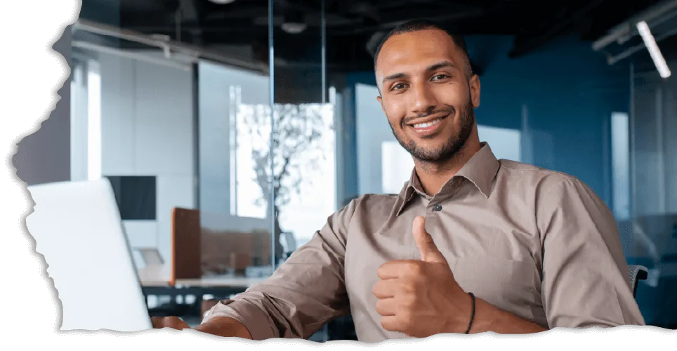Smiling man in a brown shirt giving a thumbs-up, seated at a desk in a modern office with glass walls.