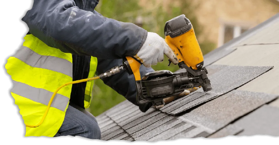 A worker in a high-visibility vest uses a nail gun to install shingles on a sloped roof. The scene conveys a focus on construction and craftsmanship.