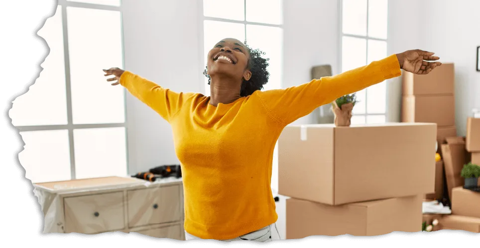 A joyful person in a yellow sweater stands with arms outstretched, smiling upward in a room filled with moving boxes. The atmosphere is light and optimistic.