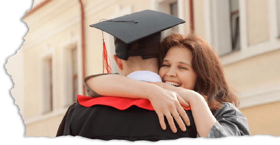 A joyful woman embraces a graduate wearing a cap and gown with a red sash, standing outside a building. The mood is celebratory and proud.