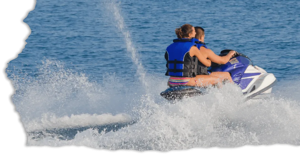A couple rides a jet ski on a sunny day, wearing blue life jackets. They create splashes and waves, conveying excitement and adventure on open water.