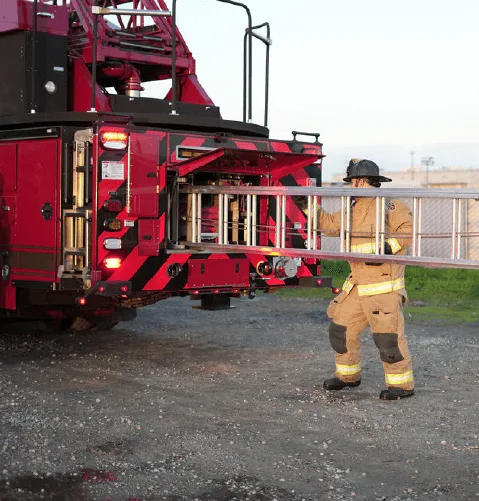 A firefighter in protective gear pulls a ladder from a red fire truck on a gravel path, showcasing readiness and duty-focused determination.