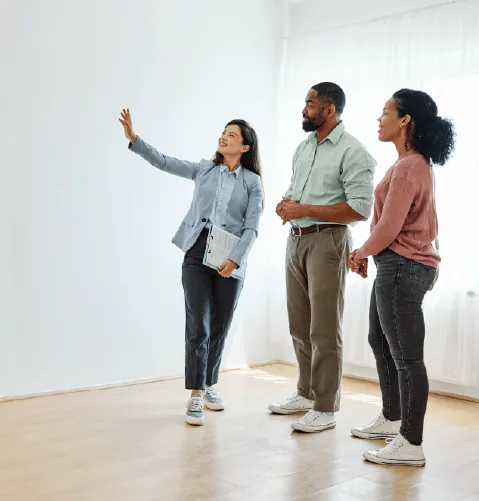 A real estate agent shows a bright, empty room to an interested couple. The agent gestures toward a wall, while the couple observes attentively.