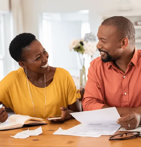 Smiling couple sitting at a table, reviewing documents. The woman in a yellow shirt holds a phone, and the man in a red shirt holds papers.