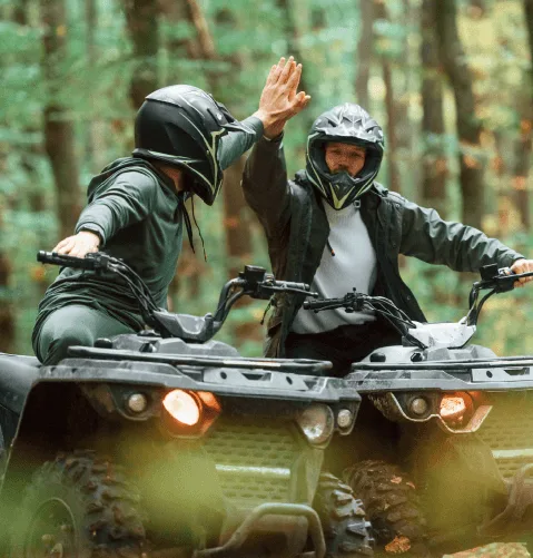 Two people wearing helmets high-five while sitting on ATVs in a forest.