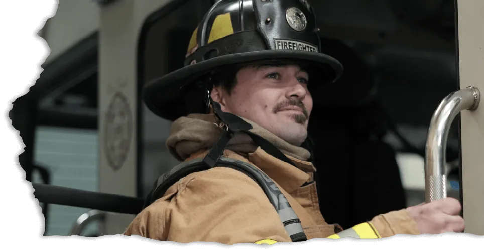 A firefighter in uniform with a helmet and yellow striped coat smiling confidently while holding onto a vehicle's handrail, conveying readiness and pride.