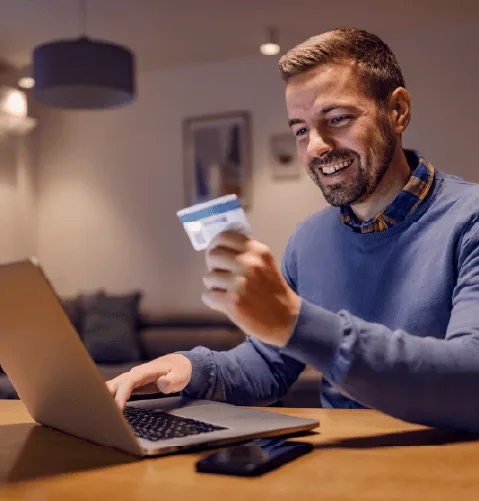 Man smiling while holding a credit card in front of a laptop. He appears to be shopping online in a cozy, dimly-lit room with a relaxed atmosphere.