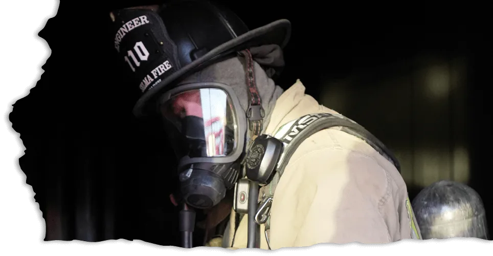 Firefighter wearing a helmet and protective gear, including a breathing mask, stands in a dimly lit space.