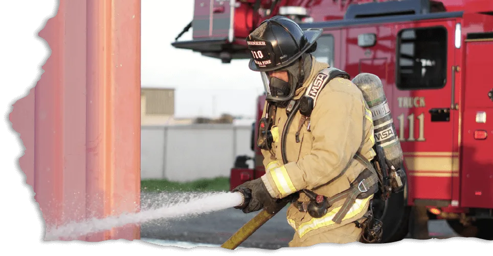 A firefighter in full gear sprays water from a hose, combating a fire. A red fire truck is visible in the background, adding urgency and bravery.