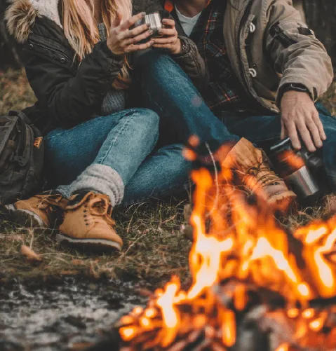 A couple sitting by a campfire, wearing warm jackets and boots. They're holding mugs, conveying warmth and coziness in a rustic outdoor setting.