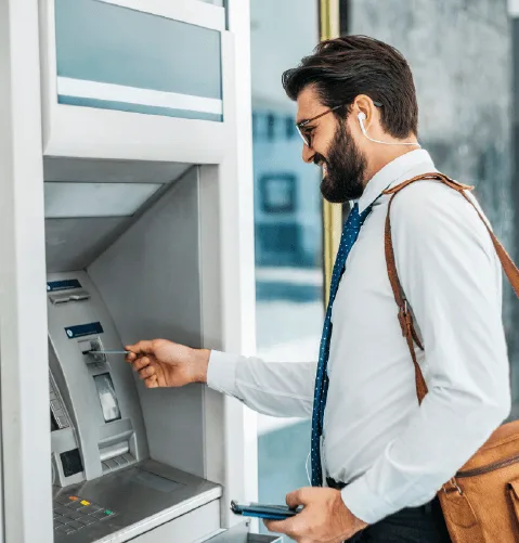 A man in a white shirt and tie uses an ATM, inserting a card. He wears earbuds and a backpack, smiling, suggesting ease and modernity.