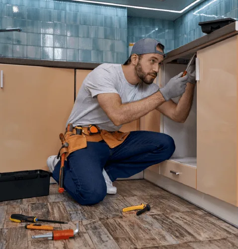 A focused man in a cap and gloves repairs a kitchen cabinet. Tools scattered on the tiled floor convey a busy, work-in-progress atmosphere.