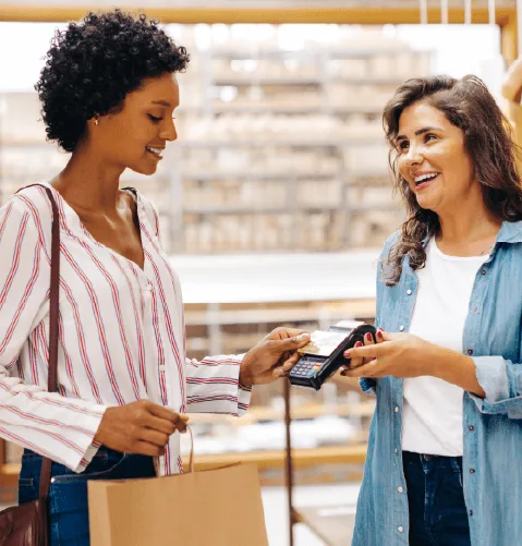 Two women smiling during a purchase. One holds a shopping bag and card, the other a payment terminal.