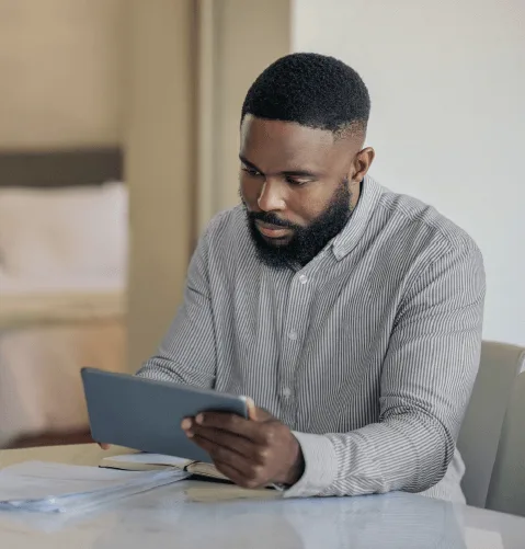 A man with a beard sits at a table, focused on a tablet. He's in a softly lit room with a bed in the background, wearing a striped shirt.