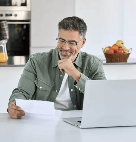 A man with glasses smiles while reading a document at a table, next to a laptop. A basket of fruit is in the background, adding warmth to the scene.