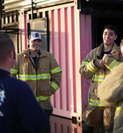 Firefighters in tan gear with reflective stripes stand together, smiling and chatting against a pink striped backdrop, conveying camaraderie and warmth.