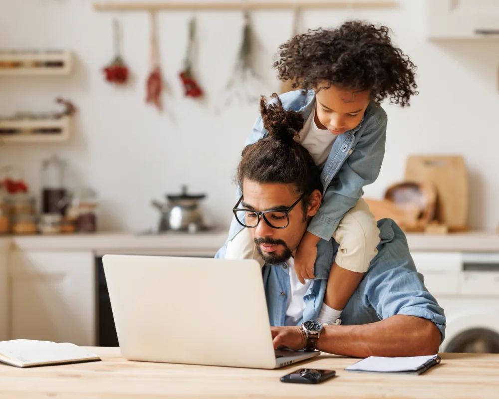 Father with child on his shoulders working on a laptop at a kitchen counter